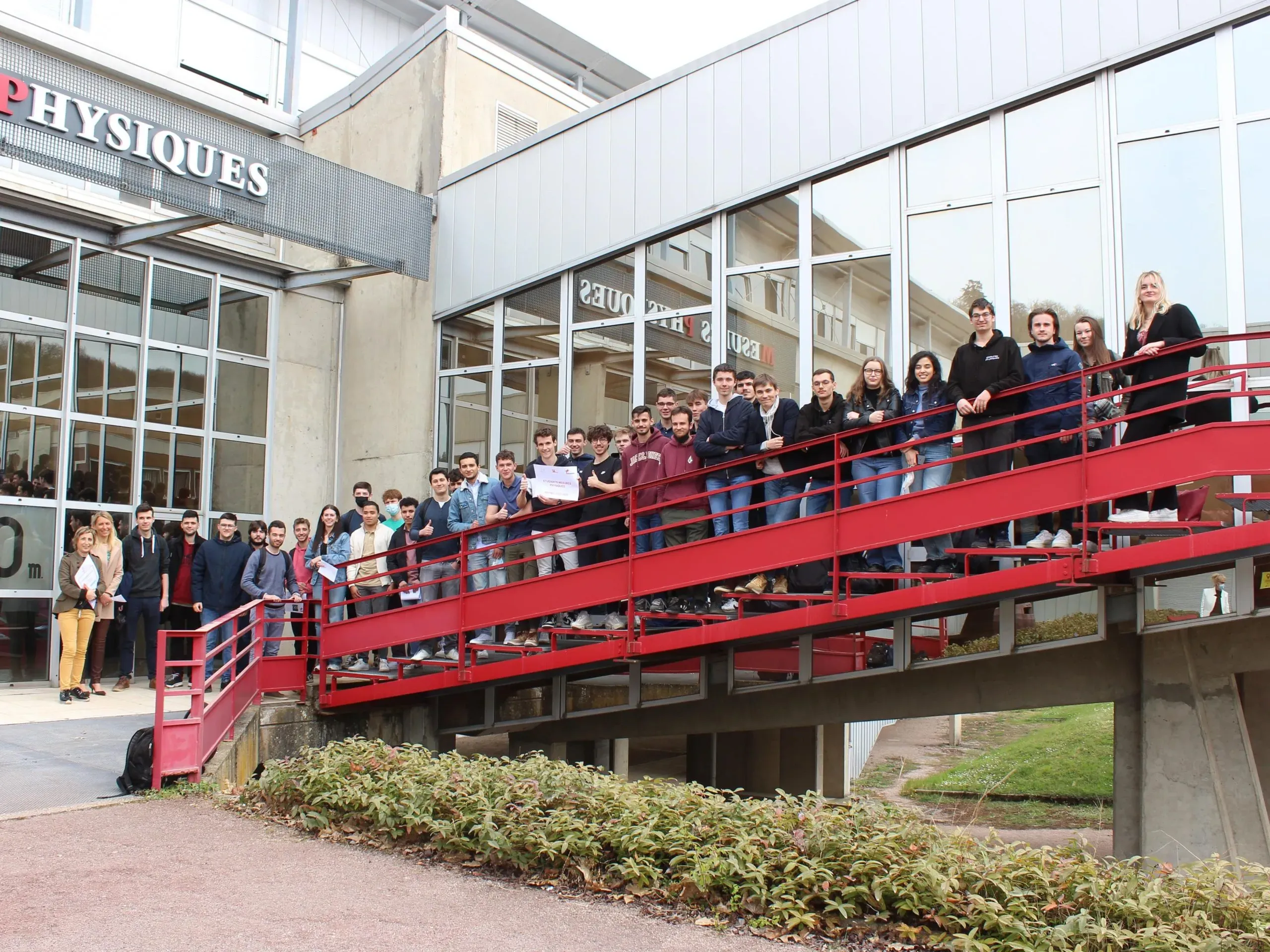 Une étudiante souriante tient un carnet de notes à la main en extérieur avec d'autres étudiants flous en arrière-plan